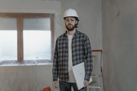 A beared man with a checked shirt and hard white hat holds a drawing pad under his arm